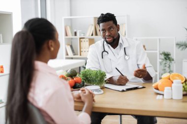 Laboratuvar önlüklü çok ırklı bir adamın seçmeli odak noktası hastane danışma odasında kadınla birlikte oturuyor. Beslenme konusunda uzman. Sağlıklı besin tercihleri konusunda tavsiyelerde bulunuyor..