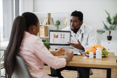 Özel poliklinikte uzun at kuyruklu bir kadın hasta. Diz üstü bilgisayardaki grafikleri kullanarak beyaz tıp üniforması ve steteskop üzerinde deneyimli gıda uzmanı tedavi etkinliğini analiz ediyor..