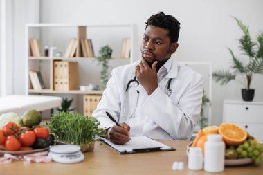 Hastanedeki çalışma saatlerinde hasta için özel bir menü hazırlama ve izin verilen ürünleri pano kağıdına yazma konusunda uzman. Afrika kökenli Amerikalı bir adam çenesine dokunuyor ve düşünüyor..