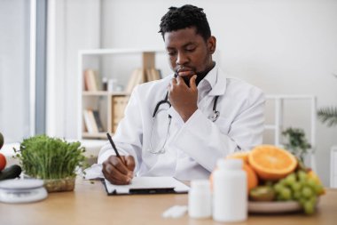 Hastanedeki çalışma saatlerinde hasta için özel bir menü hazırlama ve izin verilen ürünleri pano kağıdına yazma konusunda uzman. Afrika kökenli Amerikalı bir adam çenesine dokunuyor ve düşünüyor..