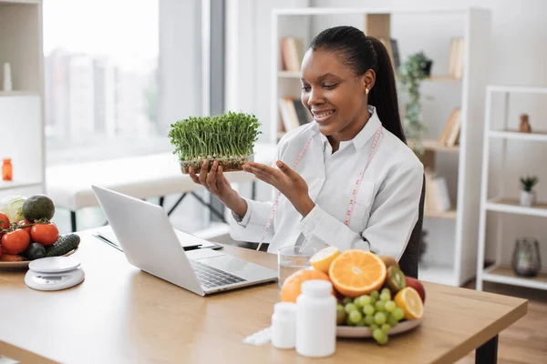 ラップトップのウェブカメラを介してクライアントとのオンライン会話中にマイクログリーンを導入アフリカ系アメリカ人女性笑顔 現代の栄養専門家健康的な食事のための若い野菜の芽を助言 — ストック写真