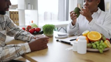 Beyaz önlüklü Afro-Amerikan diyetçisinin hasta randevusu sırasında taze avokadoyu beslenme ölçeğine koyması. Kadın profesyonel kalorilerin doğru ölçümlerini alıyor.