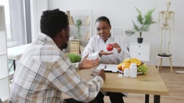 Hastane muayenesi sırasında, doktor önlüklü, taze elma tartmalı bir kadın. Çok ırklı beslenme uzmanı düşük kalorili ürünlerle beslenme düzenini geliştirme konusunda tavsiye veriyor.