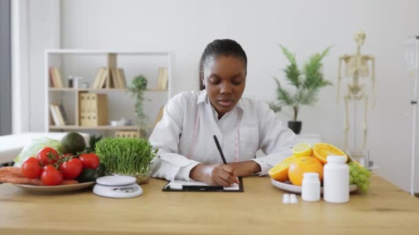 非洲裔美国女人穿着实验室外套 坐在写字台前 手握剪贴板上方的肖像 舒适工作环境下营养专业人员对患者饮食计划的改变 — 图库视频影像
