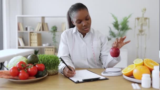コンサルティングルームで食事の尺度でリンゴを評価しながらノートを取りながら 研究室のコートで多人種女性に焦点を当てました 栄養を楽しく保つための代替を提供する思考の健康の専門家 — ストック動画