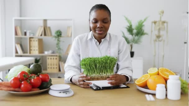 近景迷人的女士穿着白衣 拿着装有微绿色的容器 坐在办公桌前工作 非洲裔美国人营养专家建议在饮食中加入现代营养元素 — 图库视频影像