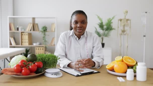 在咨询室写字台前 多文化女士与一杯清水合影的肖像 食物方面快乐的专家强调水分平衡对健康饮食的重要性 — 图库视频影像