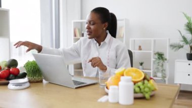 Laboratuvar önlüklü çekici yetişkin bayan kliniğin profesyonel ofisinde bilgisayarın ön kamerasına avokado gösteriyor. İnternette, meyvelerin faydaları hakkında beslenme danışmanlığı yapan çok kültürlü bir uzman..