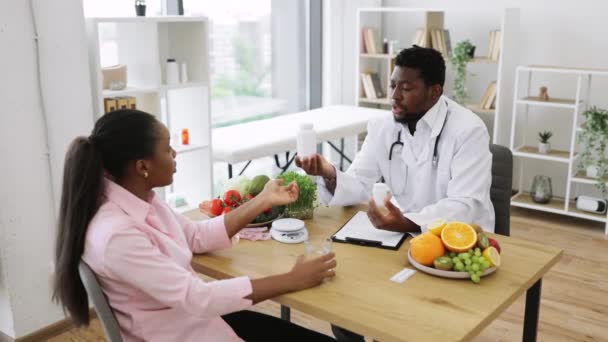 白いコートのハンサムなアフリカ系アメリカ人男性は 病院のコンサルティングルームで魅力的な女性に薬瓶を与えます 栄養専門家は栄養補助食品で全体的な健康を向上させる — ストック動画