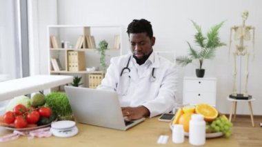 Beyaz üniformalı ve steteskoplu erkek beslenme uzmanının portresi taşınabilir bilgisayarlı ve kameraya bakıyor. Sağlıklı beslenme programı üzerinde çalışan nitelikli bir doktor..