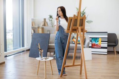 Stüdyoda kalırken elinde fırça ve palet tutan büyük beden tulumlu güzel bayanın seçici odağı. Çekici genç bir kadın tuvalde yeni elementler tanımlıyor. Doğrudan ışıklandırma kullanarak yağlı boyalarla..