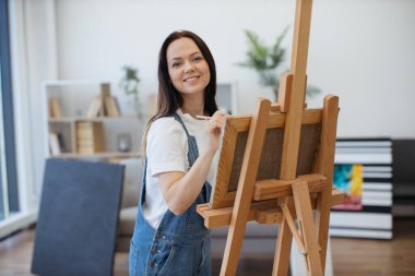 Beyaz tenli çekici bir kadın tuvale fırça darbeleri uyguluyor. İçerideki ahşap sehpa ile destekleniyor. Serbest çalışan ressam, resim stüdyosunun rahatlığını kullanarak ışık ve gölge oyunlarına konsantre oluyor..