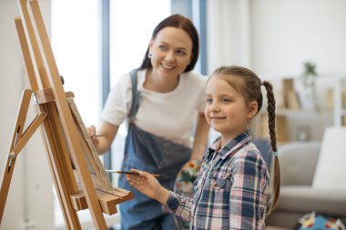 Stüdyonun içinde fırçayla tuvale boya süren güzel bir kadın ve kızın yakın görüntüsü. Neşeli sanatçı çocuklara kumaş üzerinde rehberlik ederken eğlenceli ergenlik şekilleri öğretiyor..