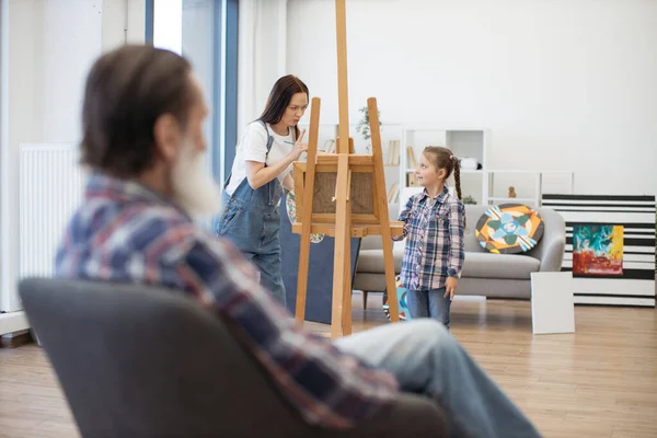 Focus on brunette lady asking cute girl about being cautious while drawing aged mans face on canvas in home workshop. Mindful parent attracting attention of daughter to details of grandpas sketch.