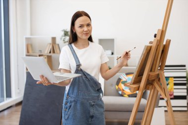 Neşeli bir kadın, kablosuz bilgisayarda grup video görüşmesi yapıyor ve arkadaşları karşılamak için geniş bir odada el sallıyor. Genç sanatçılar için online okula katılan güzel bir esmer..