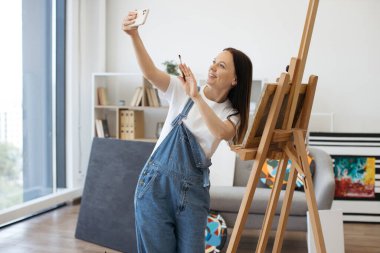 Her gün giydiği kıyafetlerle modern akıllı telefondan selfie yapan ve yaratıcı sanat stüdyosunda ahşap sehpanın yanında el sallayan olumlu bir kadın. Neşeli kadın hafta sonunu en sevdiği hobiyi yaparak geçiriyor..