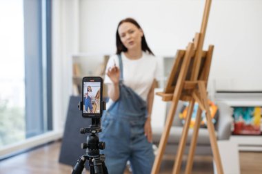 Profesyonel kadın sanatçı, kendi takipçileri için resim yaparken kişisel akıllı telefonuyla video kaydediyor. Kot tulumlu kadın, arka plan bulanık blogunda hobileri ve sanatı anlatıyor..