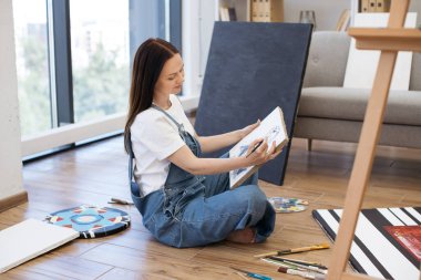 Kot pantolon giyen, ahşap zeminde oturan ve kendi sanat stüdyosunda yeni çizimler üzerinde çalışan odaklanmış kadın ressamın yan görüntüsü. Genç bayan tuvalle ve ellerinde boya fırçasıyla boş vakit geçiriyor..