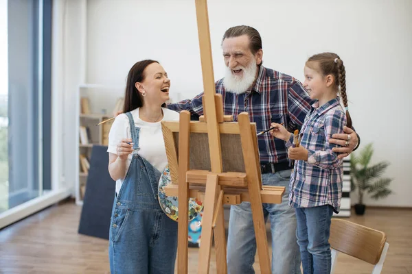 Happy ladies and senior man laughing at image on canvas while standing behind easel in home art space. Smiling mother taking break from serious painting when having fun in studio with dad and kid.