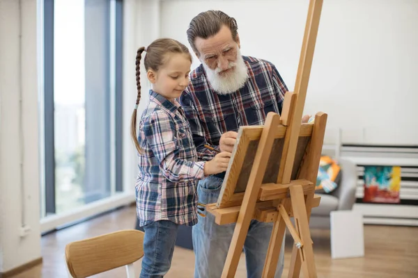 Grey-haired man preparing paint on palette while stylish girl standing in his hug. Relaxed child in denim outfit taking pleasure of art making with grandpa in studio at home.