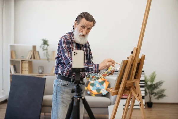 Aged man on retirement in casual outfit painting with brush on canvas using easel and looking straight at camera on smartphone set on tripod. Talented artist shooting video content about art.