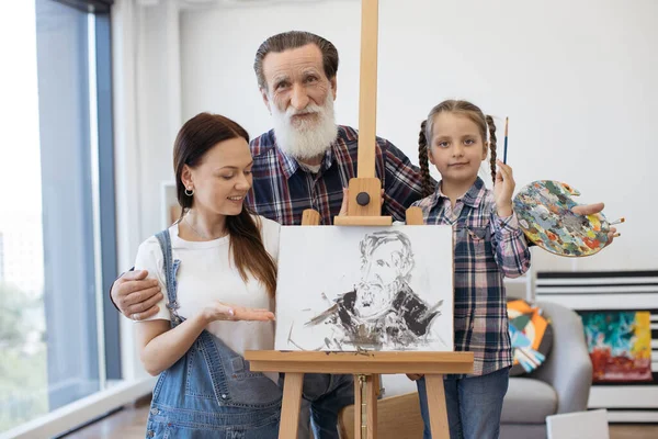 Artists family embracing together near easel with picture at bright art gallery. Grandfather with granddaughter in checkered shirts looking straight in camera while mother pointing at mans portrait.