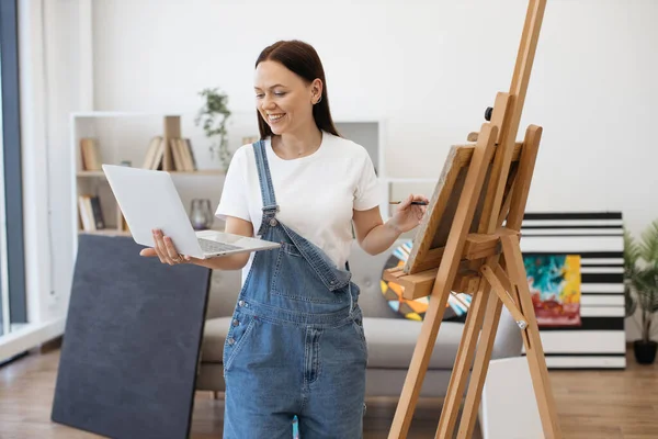 stock image Cheerful woman having group video call on wireless computer and waving hand to greet friends in bright spacious room. Pretty brunette participating in online school for young artists.