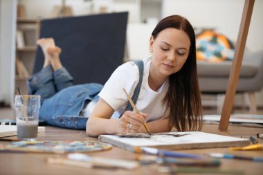 Evdeki lamine döşeme üzerinde midenin üzerine uzanmış esmer kadın resmi çiziyor. Gülümseyen kadın sanatçı sanat eseri yapıyor ve fırçayla boş zaman geçiriyor..