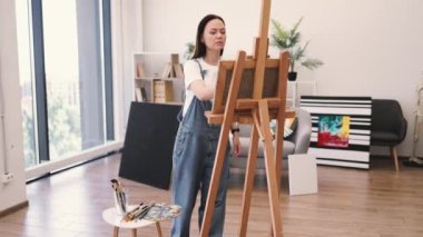 Çıplak ayaklı kadın, güneşli bir günde geniş bir sanat stüdyosunda sehpada çalışıyor. Rahat iç mekanda geniş fırça kullanarak boya uygulamasına odaklanan kendinden emin yetişkin.