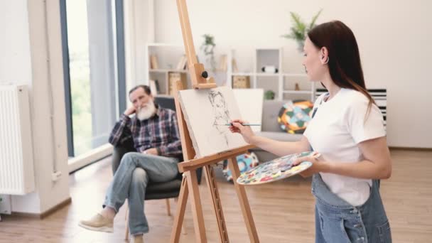現代のスタジオスペースでシッターヘッドと肩を描いたデニムオーバーオールの自営女性画家 魅力的なブルネット女性がキャンバス上のシニアマンションの肖像画の荒いアイデアをスケッチ — ストック動画