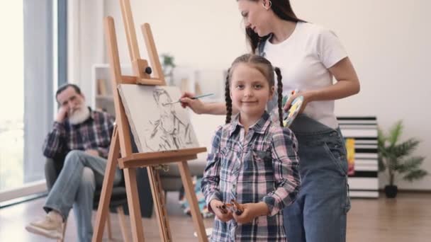 背景に座っている父親から絵を描いている間 カメラを見ている絵筆を持つ美しい少女 才能ある母親は 子供が自宅のスタジオで芸術的なプロセスに参加することを奨励 — ストック動画