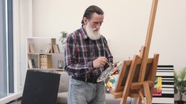 Düşünceli, gündelik giysiler içinde yaşlı bir yetişkin sanat eserlerinin kumaşında renk kombinasyonunu kontrol ediyor. Deneyimli erkek ressam tuvale doku ekleyerek belli bir görsel tasarıma yaklaşıyor..
