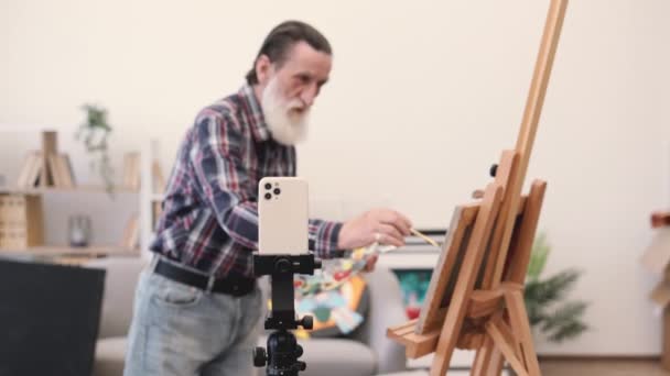 家族のアーカイブのためのビデオを撮影するための三脚の携帯電話を使用して長い灰色のひげを持つ高齢男性の画家 ジーンズとチェッカーのシャツを着て カメラのデッサンのスキルを実証したシニア男性 — ストック動画
