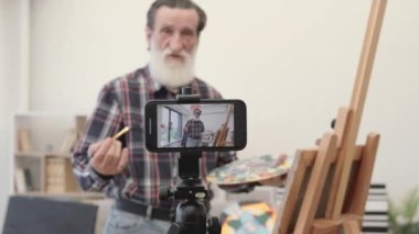 Samimi sanat stüdyosundaki resim sehpasına yeni güzel bir resim çizerken blogcu tarafından yayınlanan bulanık görüntü. Kot pantolon ve damalı gömlek giymiş yaşlı bir erkek internette takipçilerle iletişim kuruyor..