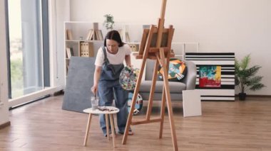 Çıplak ayaklı kadın, güneşli bir günde geniş bir sanat stüdyosunda sehpada çalışıyor. Rahat iç mekanda geniş fırça kullanarak boya uygulamasına odaklanan kendinden emin yetişkin.
