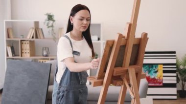 Çekici bir bayanın paletteki boyaları fırçayla karıştırırken stüdyonun içindeki kameraya gülümsemesini yakından izle. Kot tulumlar içindeki neşeli sanat eseri görsel denge için doku ekliyor.