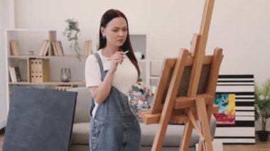 Çıplak ayaklı kadın, güneşli bir günde geniş bir sanat stüdyosunda sehpada çalışıyor. Rahat iç mekanda geniş fırça kullanarak boya uygulamasına odaklanan kendinden emin yetişkin.