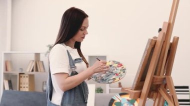 Stüdyoda kalırken elinde fırça ve palet tutan büyük beden tulumlu güzel bayanın seçici odağı. Çekici genç bir kadın tuvalde yeni elementler tanımlıyor. Doğrudan ışıklandırma kullanarak yağlı boyalarla..