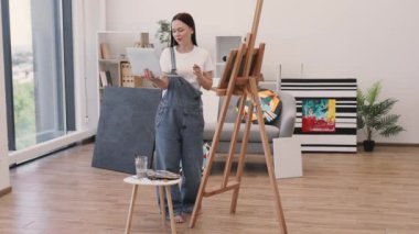 Kadın profesyonel ressam, portatif bilgisayarla video konferansı yapıyor ve panoramik manzaralı rahat bir daire kullanıyor. Yeni başlayanlarla çevrimiçi resim dersi alan günlük kıyafetli çekici bir kadın..