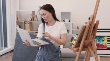 Neşeli bir kadın, kablosuz bilgisayarda grup video görüşmesi yapıyor ve arkadaşları karşılamak için geniş bir odada el sallıyor. Genç sanatçılar için online okula katılan güzel bir esmer..