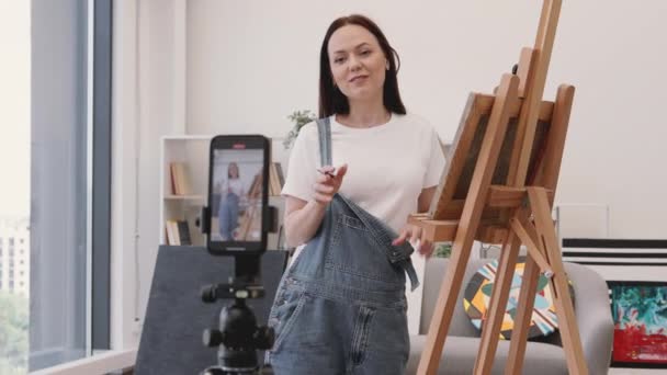 才能ある女性アーティストは アートコースを開催しながら 独自の購読者向けにオンライン放送を行うために携帯電話で三脚を使用しています 広々としたリビングルームのイーゼル近郊に立っている魅力的な長髪ブルネット — ストック動画