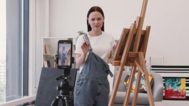 Yetenekli kadın sanatçı cep telefonlu tripodu kullanarak kendi aboneleri için online yayın yapıyor ve sanat dersleri alıyor. Büyüleyici uzun saçlı esmer, geniş oturma odasında sehpanın yanında duruyor..