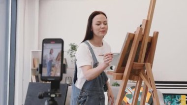 Yetenekli kadın sanatçı cep telefonlu tripodu kullanarak kendi aboneleri için online yayın yapıyor ve sanat dersleri alıyor. Büyüleyici uzun saçlı esmer, geniş oturma odasında sehpanın yanında duruyor..