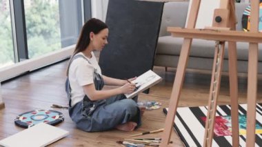 Kot pantolon giyen, ahşap zeminde oturan ve kendi sanat stüdyosunda yeni çizimler üzerinde çalışan odaklanmış kadın ressamın yan görüntüsü. Genç bayan tuvalle ve ellerinde boya fırçasıyla boş vakit geçiriyor..