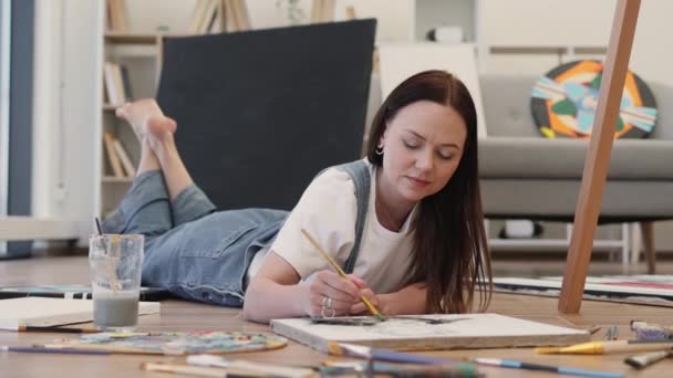 在家里躺在地板上的时候 穿着随意的黑发画着一张照片 微笑女艺术家做艺术品 用画笔打发业余时间 — 图库视频影像