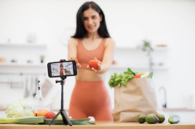 Arka planda ince bir kadın olan ve mutfak masasında tripodla tamir edilen çalışan kamera uygulaması olan akıllı cihaza odaklan. Domatesli mutlu vlogger spor besleyici gıdalarla ilgili video içeriği vuruyor.