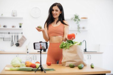 Spor giyim mağazasında elinde avokado ve tatlı biber olan neşeli bir kadın tripodda cep telefonu kullanarak online günlük yaratıyor. Atletik kadın blogcu farklı beslenme sebzelerinin faydalarını anlatıyor.