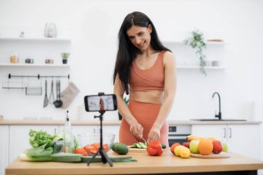 Aktif giysili gülümseyen kadın taze sebzelerden yemek hazırlıyor ve cep telefonu kamerası tripodda çalışıyor. Mutlu insan, sağlıklı beslenme geçmişi hakkında günlük türünde bir web sitesi yaratıyor..