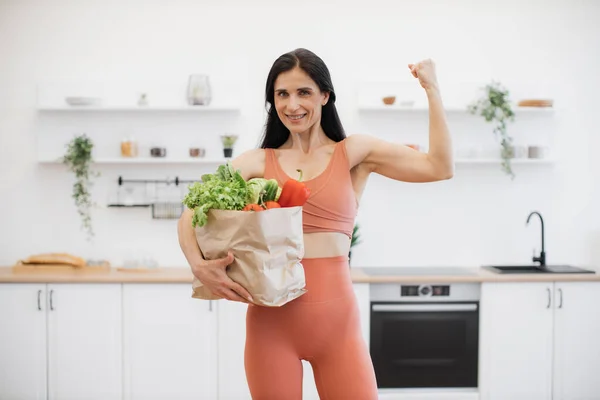 Young athletic female with package full of grocery showing arm muscles while staying at home. Slim and healthy sportswoman in gym wear promoting active lifestyle with strength in body and mind.