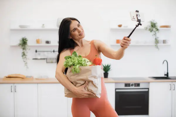 キャリアバッグを持ちながらモノポッドでモバイルカメラでポーズするアクティブウェアの陽気な女性 ホームワークアウト後の写真撮影のための新鮮な野菜と緑のスポーツブロガー — ストック写真